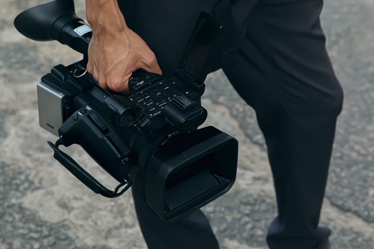 Persona sosteniendo una cámara de video profesional, apuntando hacia abajo. La cámara parece
ser de un tamaño considerable, lo que indica que puede ser utilizada para producción de video de
alta calidad. Esta imagen podría ser relevante para discusiones sobre videografía, producción de
medios o tecnología en equipos de filmación.