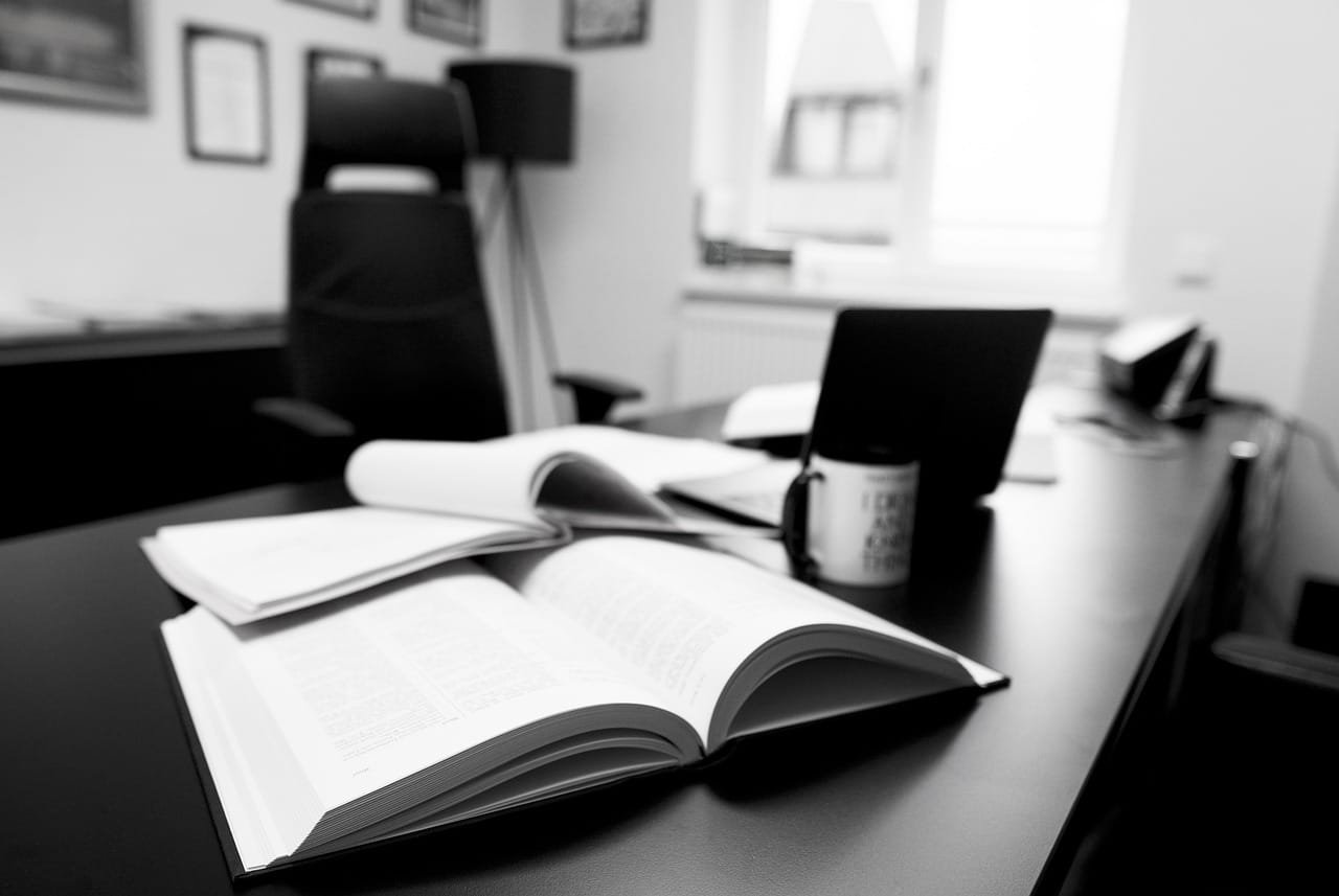Dos libros con letras borrosas colocadas uno sobre otros en una superficie plana como escritorio, junto a lo que parece ser una taza y una computadora, de fondo una silla de oficina y cuadros y diplomas de fondo un poco borrosos. Esta podría ser relevante para temas relacionados con la educación, o un ambiente de oficina .